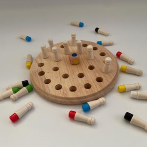 Könnte beinhalten: Holz-Steckspiel mit einer runden, hellbraunen Basis und mehreren Löchern. Das Spiel enthält kleine Holzstifte mit farbigen Spitzen, wie rot, gelb, grün, blau und schwarz. Ein kleiner Holzylinder mit gelben und blauen Seiten ist ebenfalls vorhanden.