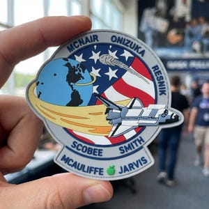 May include: A circular patch featuring a space shuttle, the Earth, and the American flag. The patch includes the names of the crew members: McNair, Onizuka, Resnik, Scobee, Smith, McAuliffe, and Jarvis. The patch has a gray border and a green apple.