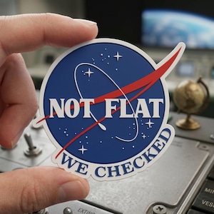 May include: A round sticker with a blue background, white text, and a red swoosh. The sticker reads "NOT FLAT" above "WE CHECKED" in a white, sans-serif font. The design is reminiscent of the NASA logo.