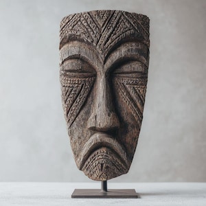 Può includere: Una maschera di legno stagionato con occhi chiusi e intricati motivi geometrici intagliati. La maschera è montata su un supporto in metallo scuro, che mostra un'opera d'arte tribale o culturale. La maschera è marrone e grigia.
