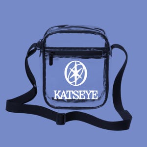 May include: Clear crossbody bag with a black adjustable strap and zipper closure. The bag features a white logo and the word "KATSEYE" printed on the front. The bag is rectangular and has a black trim.