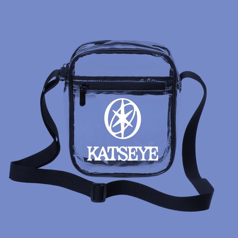 May include: Clear crossbody bag with a black adjustable strap and zipper closure. The bag features a white logo and the word "KATSEYE" printed on the front. The bag is rectangular and has a black trim.