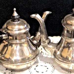 May include: Two antique silver teapots with black handles and lids. The teapots are displayed on a white lace doily against a dark background. The teapots have a classic design, with a rounded body and a spout.