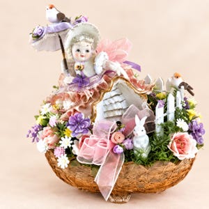 May include: A decorative figurine arrangement in a brown woven basket featuring a doll with a parasol, a small white house, a white rabbit, and two birds. The scene is decorated with pink and purple fabric flowers, green foliage, and a white picket fence.