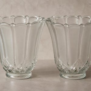 May include: Two clear glass tulip-shaped candle holders. The holders have a frosted, fluted design with a diamond pattern near the base. The rims are scalloped. The candle holders are suitable for home decor.