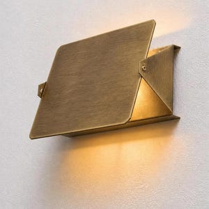 Könnte beinhalten: Moderne, geometrische Wandleuchte in warmem Messing-Finish. Die Leuchte hat ein rechteckiges Design mit einer gefalteten, abgewinkelten Oberseite und einem dreieckigen Sockel. Die Leuchte strahlt ein weiches, warmes Licht aus.