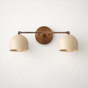 May include: A two-light wall sconce with a bronze finish. The fixture has a circular backplate, a horizontal bar, and two cream-colored, dome-shaped shades. Each shade houses a lightbulb. The sconce is mounted on a white wall.