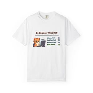 Op de afbeelding: Wit T-shirt met een cartoonkat met een bril die op een computer werkt. De tekst "QA Engineer Checklist" staat boven de kat, met een lijst met items waaronder "Life unstable" en "Build stable".