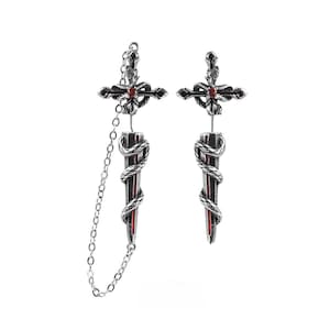 May include: A pair of silver-toned earrings featuring a dagger and cross design. Each earring has a snake coiled around the blade, which is accented with a red centre. One earring includes a delicate silver chain connecting the cross to the dagger.