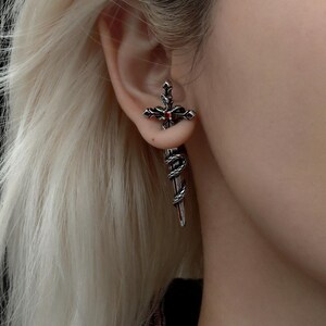 May include: A silver-toned metal earring shaped like a sword with a cross-guard. The hilt features a small, round, red gemstone at the centre. A textured, snake-like design wraps around the blade of the sword, which hangs vertically from the earlobe.