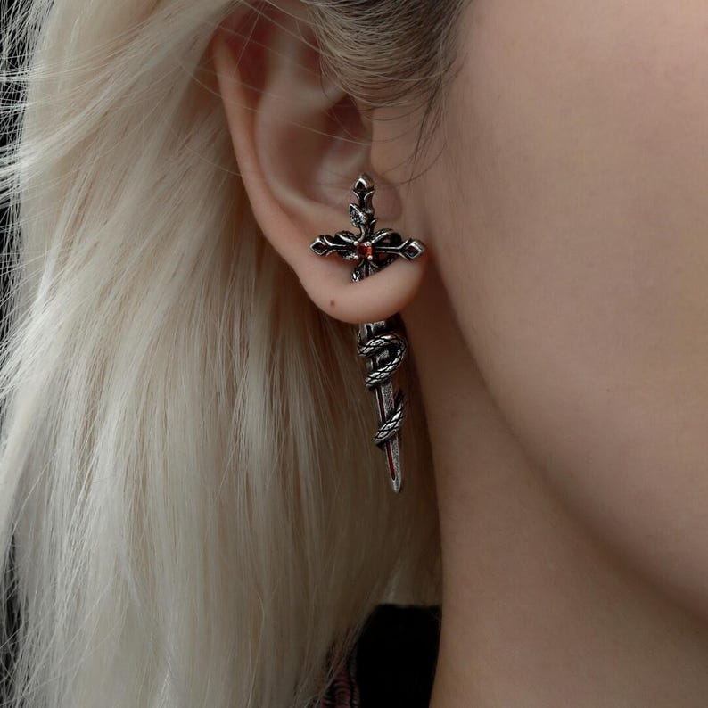 May include: A silver-toned metal earring shaped like a sword with a cross-guard. The hilt features a small, round, red gemstone at the centre. A textured, snake-like design wraps around the blade of the sword, which hangs vertically from the earlobe.