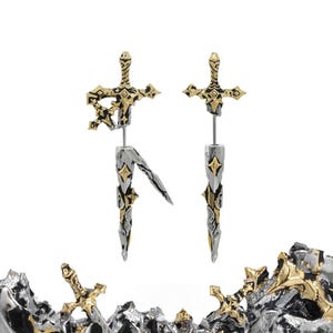 May include: A pair of earrings shaped like swords with gold and silver metallic finishes. The top section features a hilt design, while the bottom section resembles a blade. The earrings have a textured, weathered appearance with small star-shaped accents.