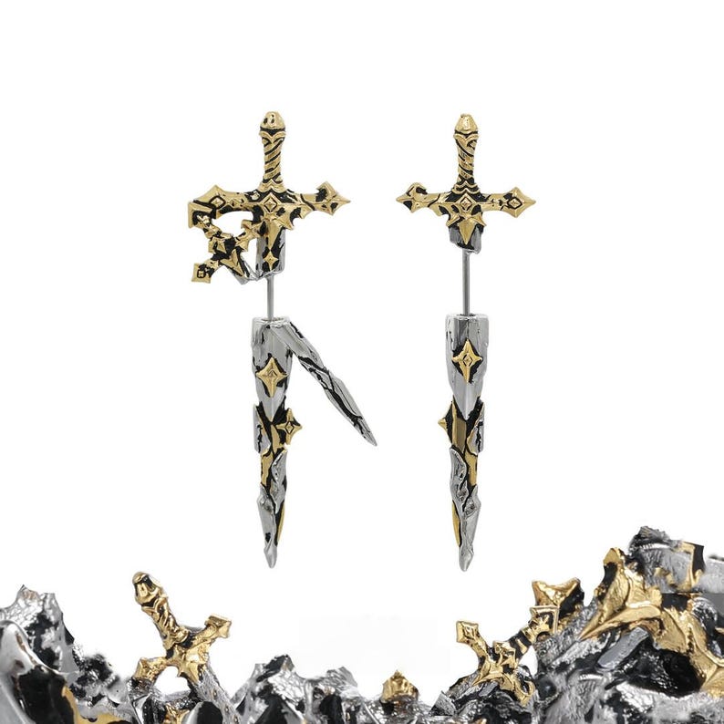 May include: A pair of earrings shaped like swords with gold and silver metallic finishes. The top section features a hilt design, while the bottom section resembles a blade. The earrings have a textured, weathered appearance with small star-shaped accents.