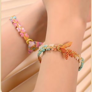May include: Two bracelets featuring small, heart-shaped fabric elements in pastel pink, blue, and orange hues, woven with a thin gold-colored thread. One bracelet includes a prominent orange fabric fish tail charm, while the other has a striped tail.