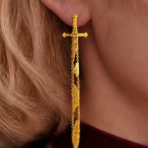 May include: A gold-toned dangle earring shaped like a sword. The earring features a sword hilt at the top, with a long, slender blade section connected by delicate gold chains. The surface of the sword is decorated with intricate, embossed patterns.