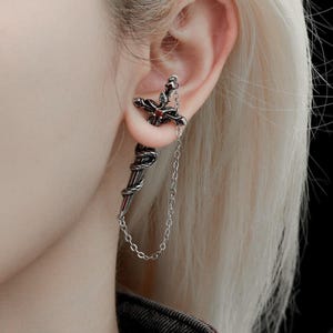 May include: A silver-toned ear cuff featuring a sword design wrapped in a snake. The sword hilt is accented with a small red stone. A delicate silver chain connects the top of the sword to the bottom tip, creating a draped effect on the earlobe.
