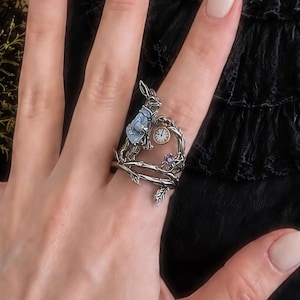 May include: A silver-toned ring featuring a rabbit wearing a light blue jacket, a small round clock with black hands, and a single purple gemstone. The band is designed to resemble intertwined tree branches with textured bark details.