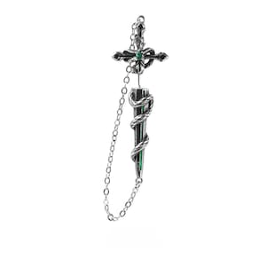 May include: A silver-toned metal earring featuring a cross-shaped top with a small green stone at the centre, connected by a delicate chain to a dagger-shaped pendant wrapped in a coiled snake, with a thin green vertical line along the blade.