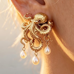 Puede incluir: Un pendiente dorado con forma de pulpo que presenta tentáculos texturizados y una cabeza lisa. Tres perlas blancas en forma de lágrima cuelgan de la parte inferior del pulpo, suspendidas por pequeños anillos dorados en los tentáculos.