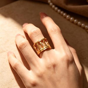 May include: A wide gold band ring featuring a series of vertical cutouts and raised, pillar-like architectural details around the circumference. The metal has a polished, reflective finish that catches the light, creating a structured, openwork design.