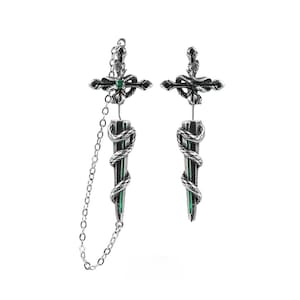May include: A pair of silver-toned earrings featuring a sword and snake motif. The top section is a cross-shaped hilt with a small green stone, while the blade is wrapped in a coiled snake. One earring includes a delicate silver chain connector.