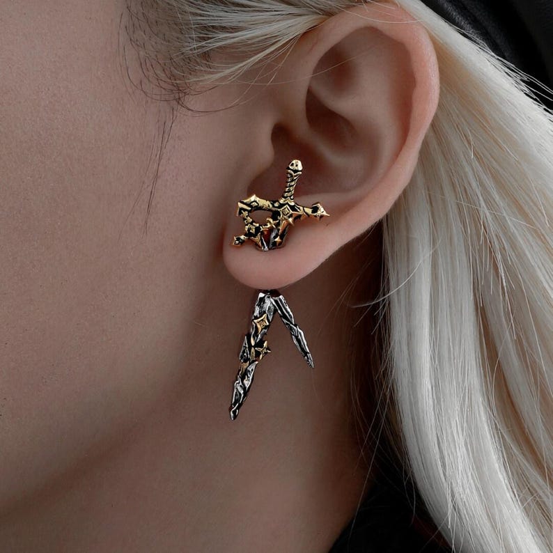 May include: A two-piece ear stud featuring a gold-toned sword hilt design that sits in the earlobe, paired with a dangling silver-toned sword blade attachment that hangs below, accented with small star-shaped details on the metal surfaces.