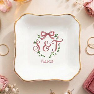 May include: A square white ceramic trinket dish with a scalloped edge and a gold-painted rim. The center features a pink floral wreath graphic with a bow, the letters S & T, and the text Est. 2026 printed in a matching pink script font.