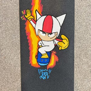 Puede incluir: Lija de monopatín negra con un personaje de dibujos animados con un casco blanco con una raya roja, montado en un monopatín azul. El personaje está rodeado de llamas amarillas, naranjas y rojas. Texto estilo grafiti en la parte inferior.