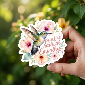 May include: A sticker featuring a hummingbird with green, blue, and orange feathers, surrounded by pink and purple hibiscus flowers. The sticker has the words "practice radical Empathy" in a script font.