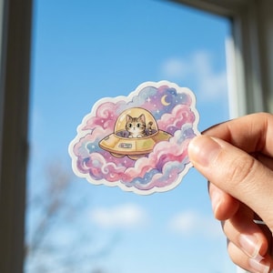 May include: A sticker featuring a cartoon cat in a yellow UFO, surrounded by pastel pink, purple, and blue clouds. A crescent moon and stars are visible in the background. The sticker is held by a hand.