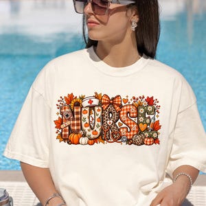 Op de afbeelding: Een off-white t-shirt met korte mouwen en een grafische print van het woord NURSE in grote blokletters. De letters zijn versierd met herfstthema-patronen, waaronder pompoenen, esdoornbladeren, zonnebloemen, een verpleegsterskapje en een stethoscoop.