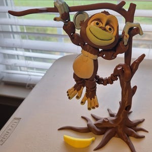 May include: A brown monkey figurine hanging from a brown tree branch. The monkey has a light brown face, a tan belly, and light blue hands and feet. A yellow banana rests on the surface below the tree.