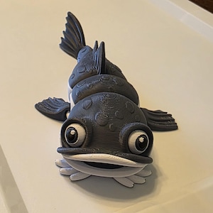 May include: A dark gray articulated fish figurine with a textured, spotted body, large white eyes with black pupils, and a wide white mouth. The fish features multiple segments along its back, side fins, and a tail fin, all in a matte finish.