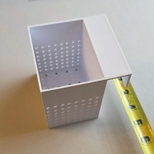 May include: A white rectangular container with a perforated design on the sides and bottom. The container has a flat top with an extended edge. A yellow measuring tape is next to the container, showing a measurement of approximately 5 inches.