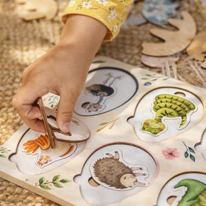 Könnte beinhalten: Holzpuzzle mit Tierillustrationen, darunter ein Strauß und eine Schlange. Eine Person setzt die Puzzleteile zusammen. Die Person trägt ein gelbes Hemd mit Blumenmuster. Das Puzzle liegt auf einer hellen Holzoberfläche.