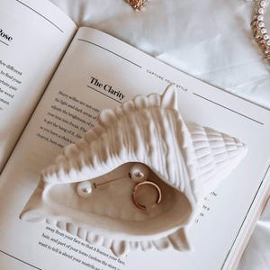 Può includere: Un portagioie bianco a forma di conchiglia contiene un anello d'oro e un orecchino di perle. Il piatto è appoggiato su un libro aperto con il titolo "The Clarity" e le parole "Capture Your Style". Sono visibili anche orecchini d'oro e una collana di perle.