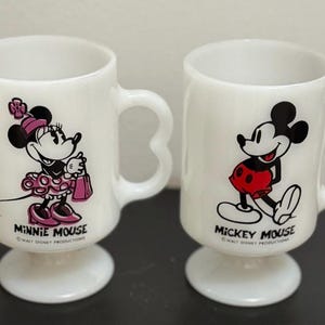 May include: Two white ceramic mugs featuring Mickey Mouse and Minnie Mouse. The mugs have heart-shaped handles and pedestal bases. Minnie Mouse is depicted in a pink dress, and Mickey Mouse is in red shorts. "Minnie Mouse" and "Mickey Mouse" are printed below the characters.