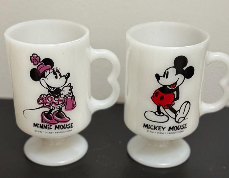May include: Two white ceramic mugs featuring Mickey Mouse and Minnie Mouse. The mugs have heart-shaped handles and pedestal bases. Minnie Mouse is depicted in a pink dress, and Mickey Mouse is in red shorts. "Minnie Mouse" and "Mickey Mouse" are printed below the characters.