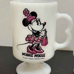 May include: White glass Minnie Mouse mug with a pedestal base and heart-shaped handle. The mug features a black and pink graphic of Minnie Mouse, holding a pink purse. The text "MINNIE MOUSE" and "WALT DISNEY PRODUCTIONS" are printed on the mug.