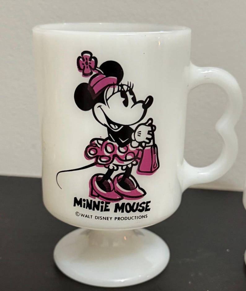May include: White glass Minnie Mouse mug with a pedestal base and heart-shaped handle. The mug features a black and pink graphic of Minnie Mouse, holding a pink purse. The text "MINNIE MOUSE" and "WALT DISNEY PRODUCTIONS" are printed on the mug.