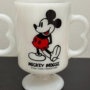May include: White Mickey Mouse mug with a heart-shaped handle. The mug features a smiling Mickey Mouse graphic in black, red, and white. The words "Mickey Mouse" and "Walt Disney Productions" are printed on the mug.