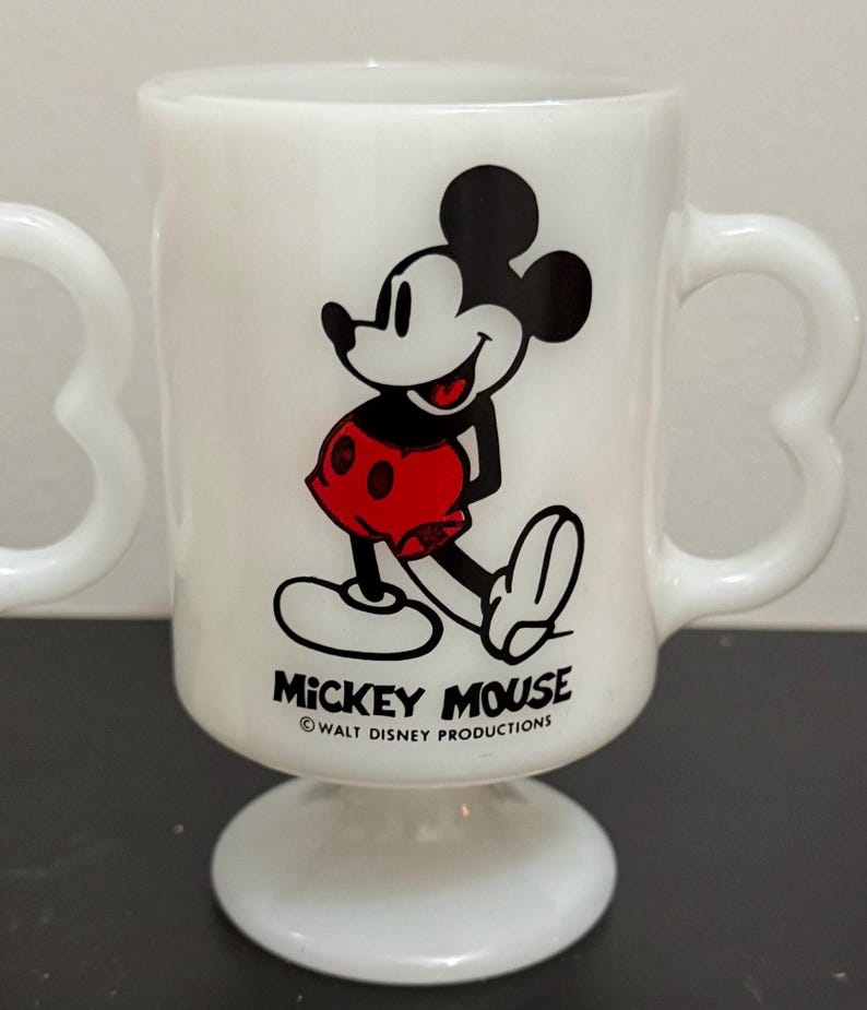 May include: White Mickey Mouse mug with a heart-shaped handle. The mug features a smiling Mickey Mouse graphic in black, red, and white. The words "Mickey Mouse" and "Walt Disney Productions" are printed on the mug.