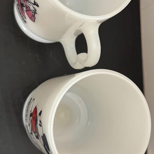 May include: Two white ceramic mugs with curved handles. Each mug features a cartoon character design, one with Minnie Mouse in pink and the other with Mickey Mouse in red. The mugs are set against a dark background.