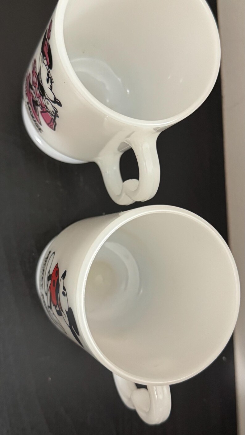 May include: Two white ceramic mugs with curved handles. Each mug features a cartoon character design, one with Minnie Mouse in pink and the other with Mickey Mouse in red. The mugs are set against a dark background.