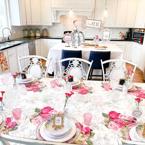 Op de afbeelding: Een witte tafel met een bloemen tafelkleed gedekt voor een feestje. Er staan roze anjers in kleine glazen vazen, gouden fotolijstjes met plaatskaarten en witte borden met gouden randen. De tafel is gedekt met heldere glazen en roze glazen bekers. De tekst op de plaatskaarten luidt "The Shop" en "Garden".