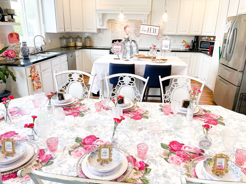 Op de afbeelding: Een witte tafel met een bloemen tafelkleed gedekt voor een feestje. Er staan roze anjers in kleine glazen vazen, gouden fotolijstjes met plaatskaarten en witte borden met gouden randen. De tafel is gedekt met heldere glazen en roze glazen bekers. De tekst op de plaatskaarten luidt "The Shop" en "Garden".