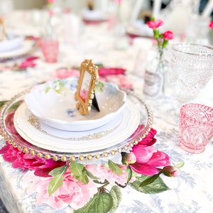 Op de afbeelding: Een tafeldekking met een wit bord, een gouden onderbord en een bloemen placemat. Een klein gouden fotolijstje staat op het bord. De tafel is bedekt met een wit tafelkleed met een bloemenpatroon.