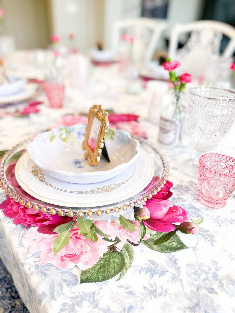 Op de afbeelding: Een tafeldekking met een wit bord, een gouden onderbord en een bloemen placemat. Een klein gouden fotolijstje staat op het bord. De tafel is bedekt met een wit tafelkleed met een bloemenpatroon.