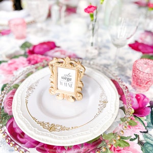 Op de afbeelding: Een wit bord met een gouden rand en een gouden decoratief frame met de tekst "votre si&egrave;ge wine" op een bloemige tafelsetting met roze bloemen.