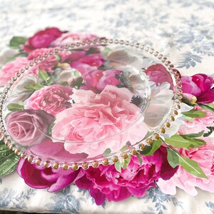Op de afbeelding: Een heldere glazen serveerschotel met een gouden parelrand ligt op een onderzetter met bloemenprint. De onderzetter heeft roze, witte en groene pioenen.